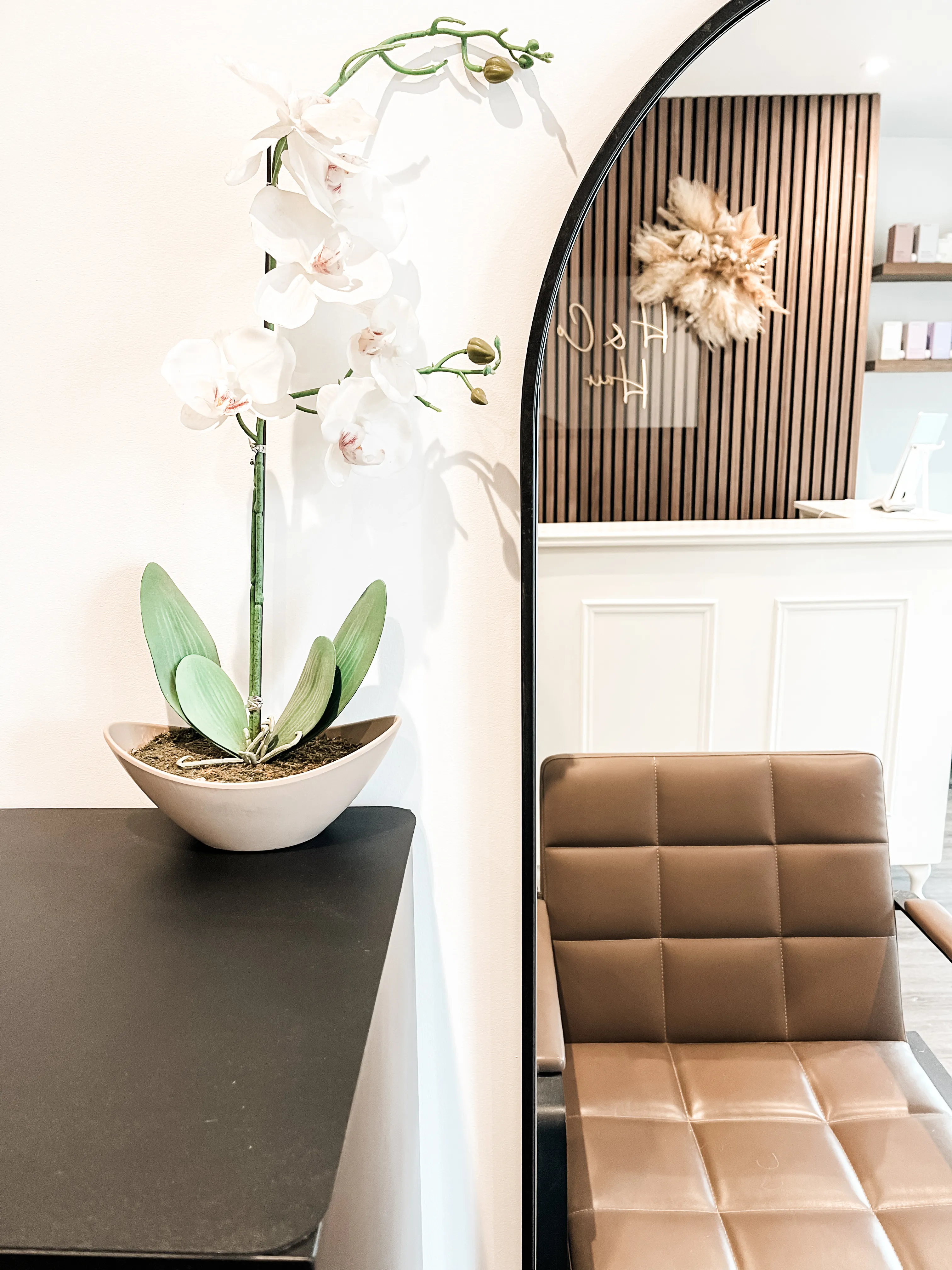 The H&Co salon reception, with a black-framed arch mirror, orchid in a stone bowl, oak slat wall and tan leather styling chair.