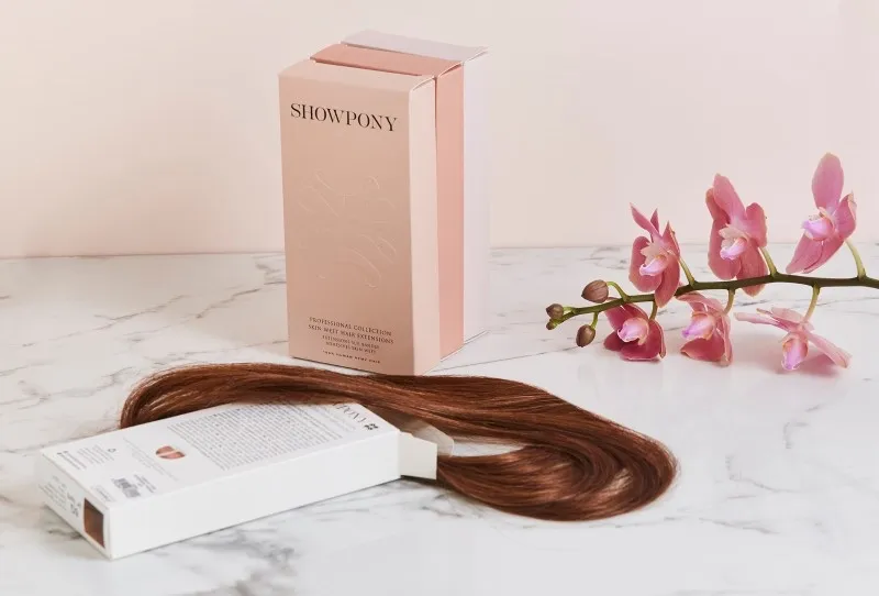 Showpony skin weft hair extensions box and a length of brunette weft on marble, with a pink orchid stem.